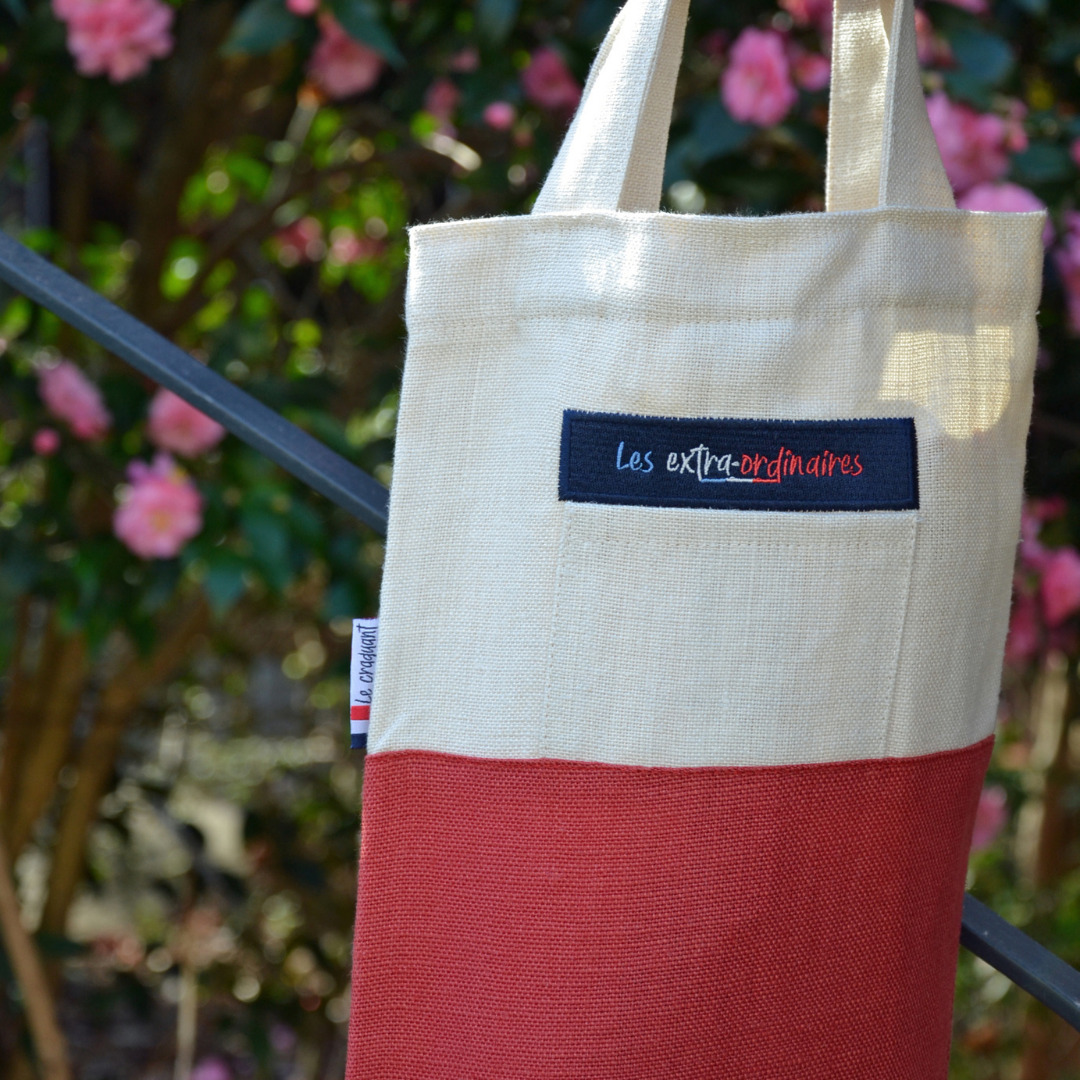 Sac à pain le craquant rouge tenu par les anses devant buisson en fleurs